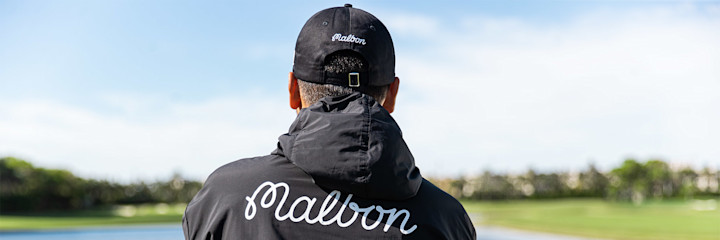 Person bakifrån bär svart keps och jacka med texten "Malbon", står utomhus vid en golfbana under klar himmel.