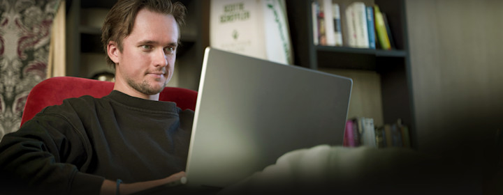 Man sitter i en röd fåtölj och arbetar på en bärbar dator i ett rum med bokhyllor i bakgrunden.