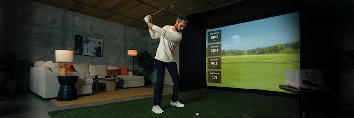 Man svingar en golfklubba framför en golfsimulator med projicerad bana och statistik i ett modernt vardagsrum.