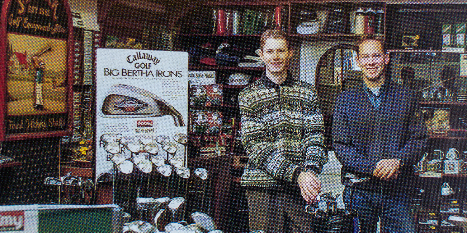 Två leende män står bredvid golfklubbor i en golfbutik med hyllor och en skylt för Callaway Big Bertha Irons i bakgrunden.