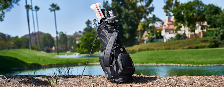 En svart golfbag med texten "GHOST" står på marken vid en golfbana, med klubbor och klubbskydd synliga, gräs och vatten i bakgrunden.