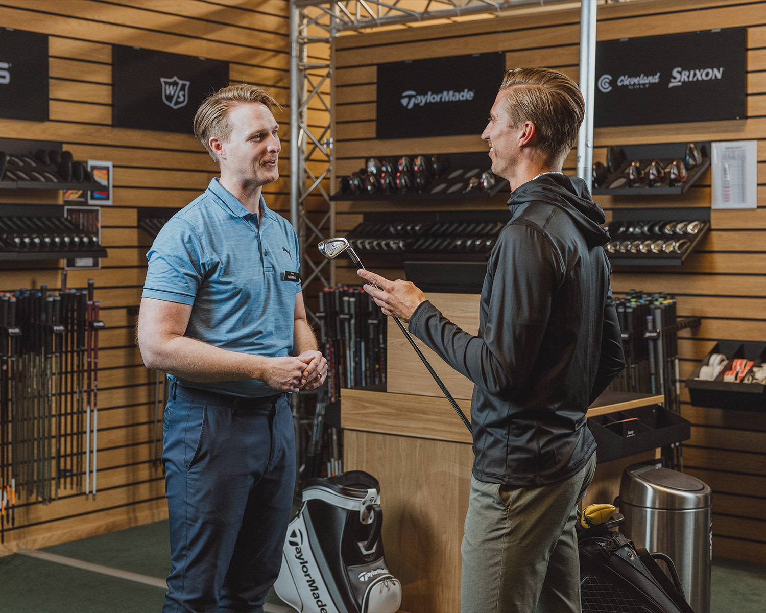 Två män samtalar i en golfbutik, där en håller en golfklubba och den andra bär butikskläder, omgiven av golfutrustning och klubbor.
