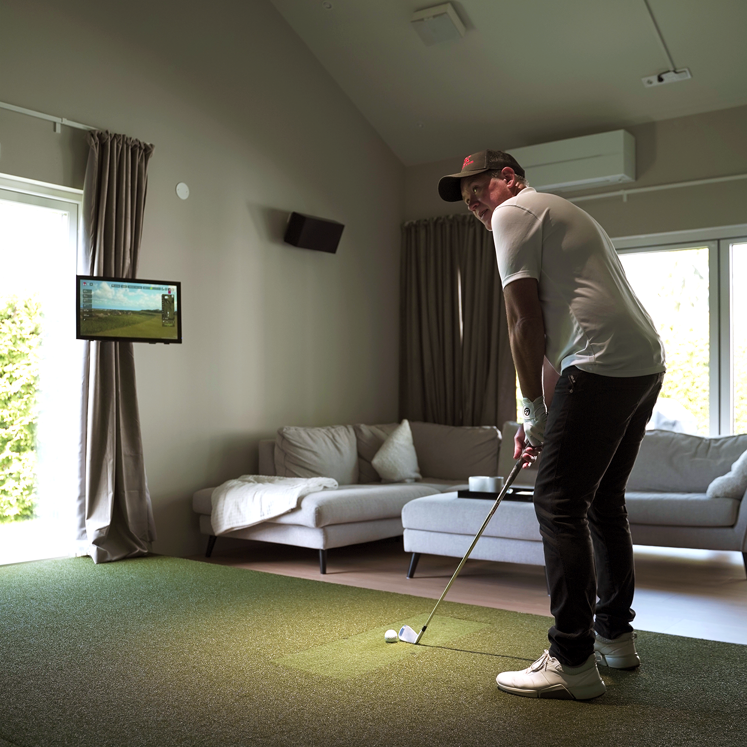 Man spelar golf med simulator i vardagsrum, står på konstgräs med klubba, soffa och TV-skärm i bakgrunden.