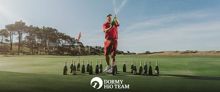 En man i röd golfklädsel öppnar en champagneflaska på en golfbana, omgiven av fler flaskor, med skog och klubbhus i bakgrunden.