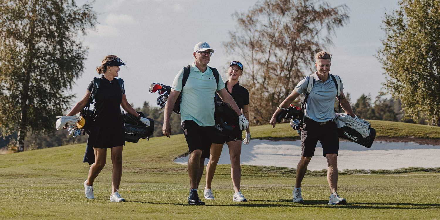 Fyra personer går tillsammans på en golfbana, bärandes golfbagar och klädda i sportkläder under en solig dag.