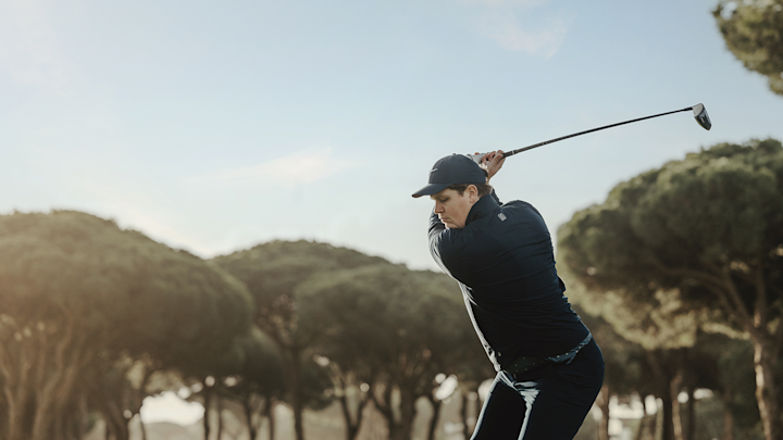 Un homme vêtu de vêtements sombres et d'une casquette balance un club de golf sur un parcours de golf, avec des arbres en arrière-plan.