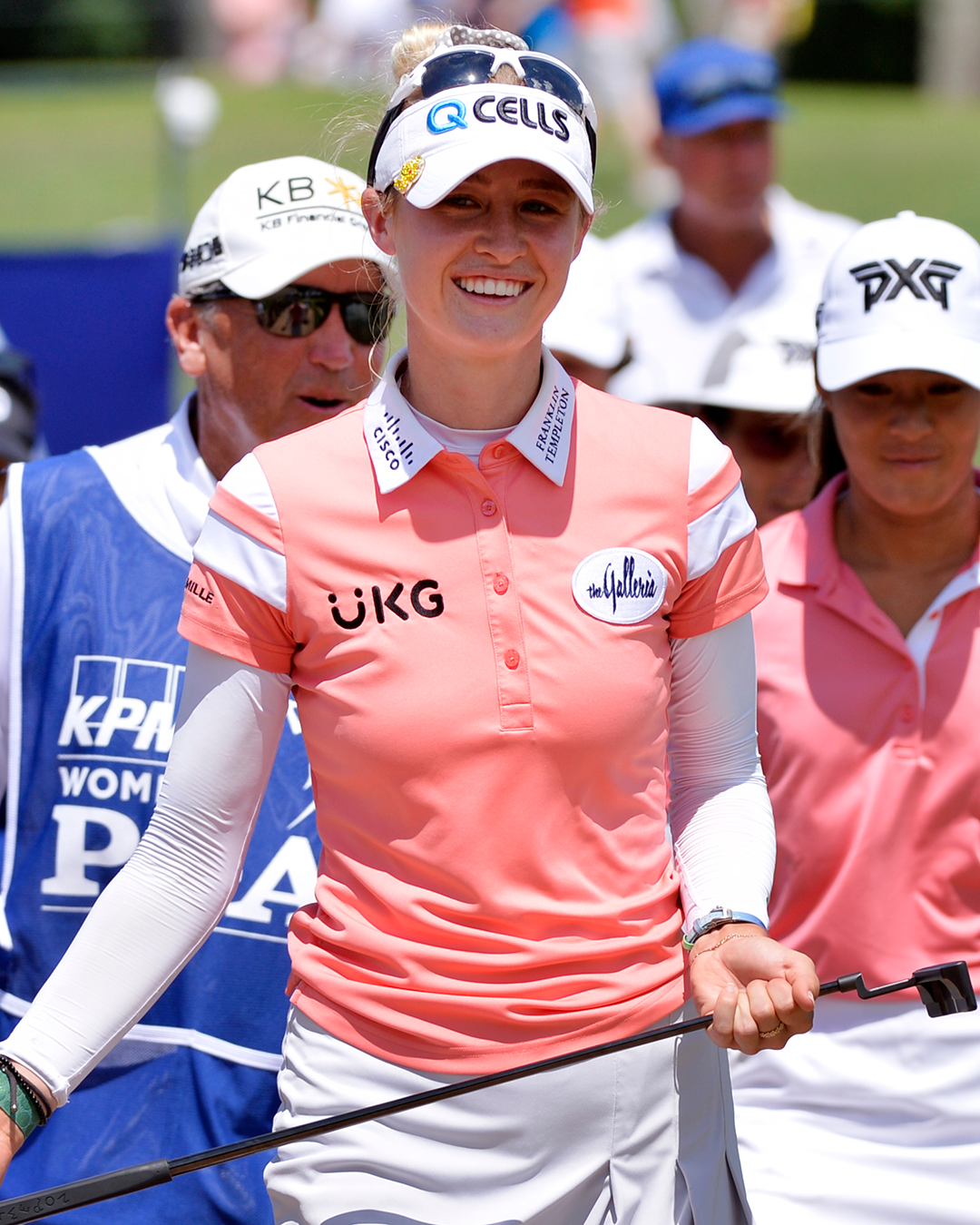 Female golfer in a pink jumper and white cap holding a golf club and smiling on the golf course, surrounded by other people.