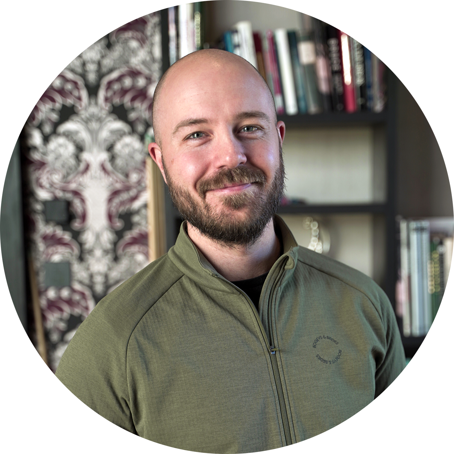 Un homme barbu à la tête rasée sourit et se tient devant une étagère remplie de livres, avec un papier peint à motifs en arrière-plan.