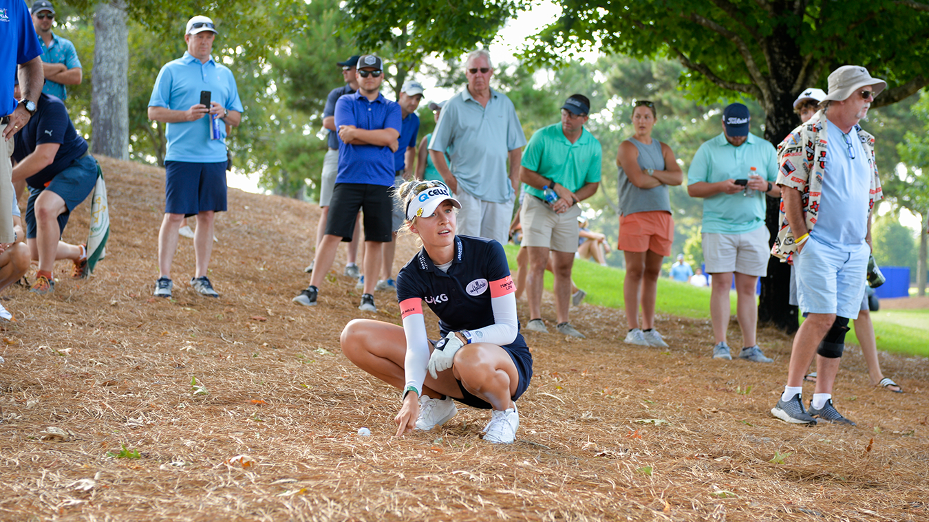 A golfer crouches down and points at a golf ball on the ground, surrounded by spectators in a relaxed outdoor setting.