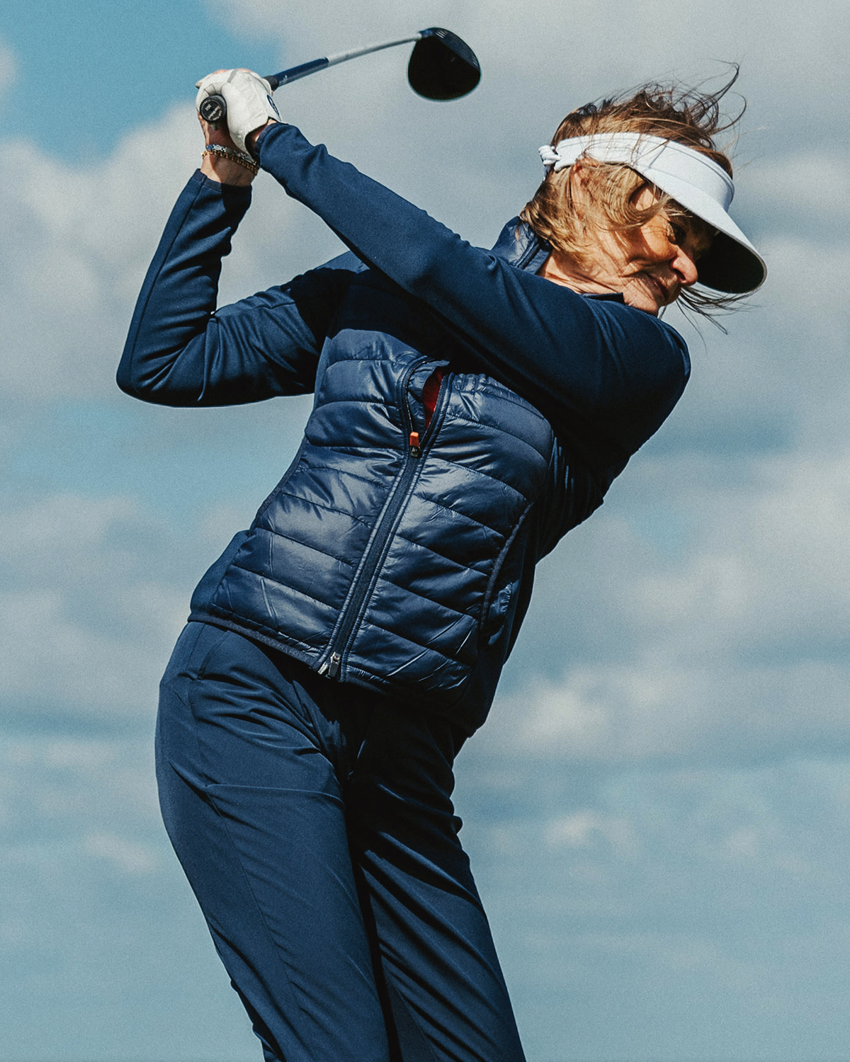 Kvinna i blå golfkläder och vit visir slår ett golfslag utomhus under en molnig himmel.