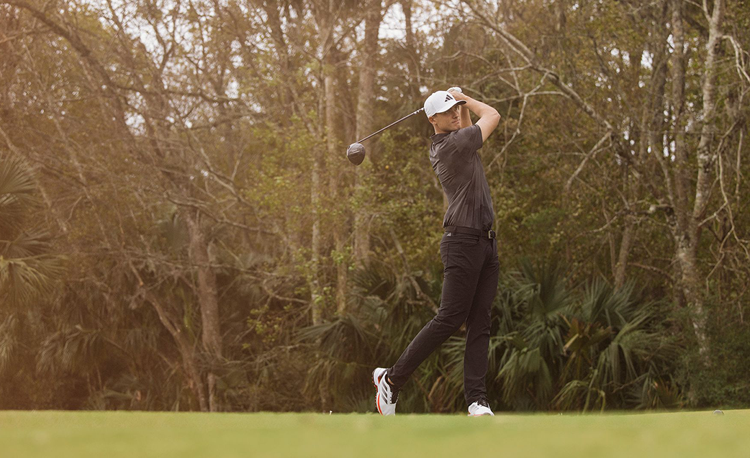 Un golfeur balance un club sur un terrain de golf verdoyant entouré d’arbres et de végétation.