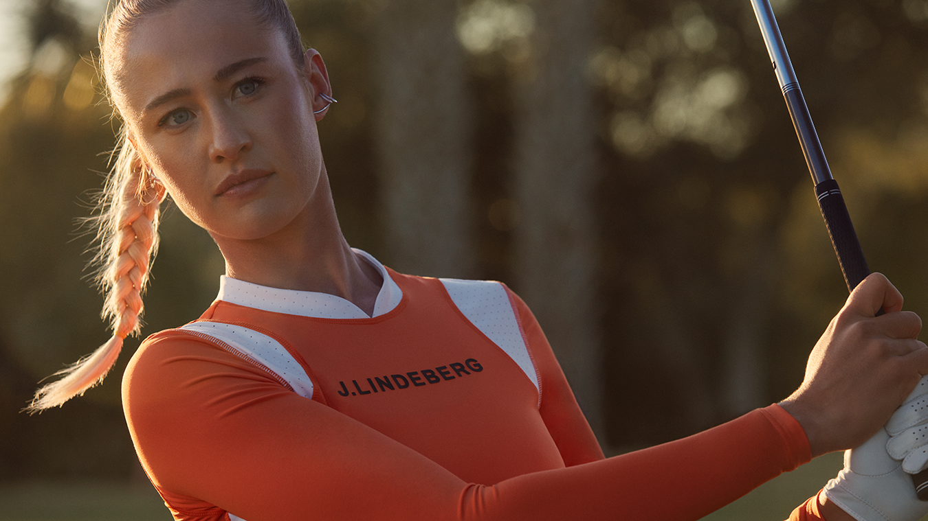 Woman with a braid holding a golf club, wearing an orange jumper with the text "J.LINDEBERG" outdoors in the sunlight.