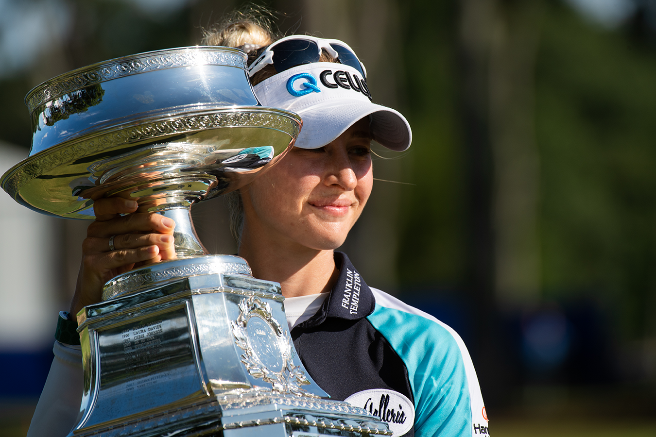 Female golfer holding up a large silver trophy and smiling outdoors on a golf course.